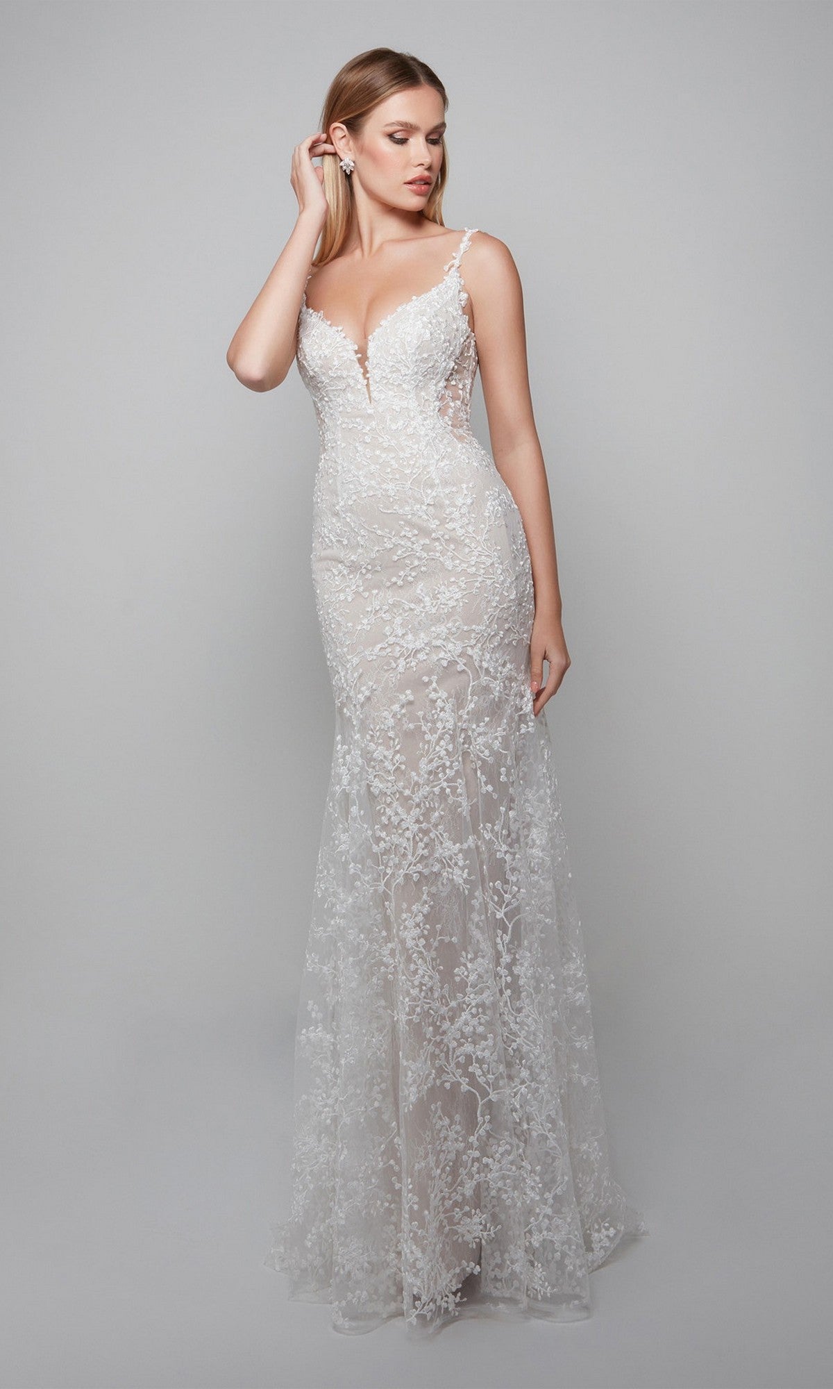 A woman poses against a light gray background wearing the Alyce 7077, a fitted floor-length ivory/lt latte dress in a trumpet silhouette with thin straps, lace appliques, and a deep V neckline.